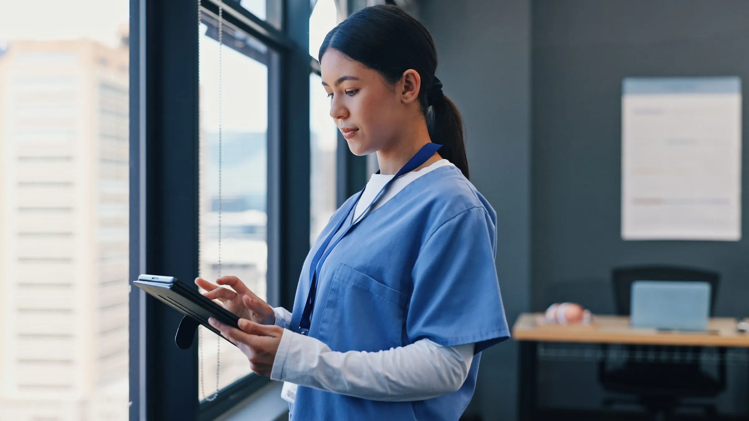 A nurse in scrubs reviewing information on a tablet beside an office window