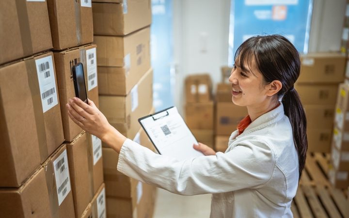 A warehouse worker in a white coat scanning boxes with a phone while holding a clipboard