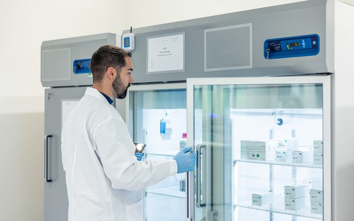 A pharmacist in a lab coat and gloves opening a temperature-controlled medicine fridge