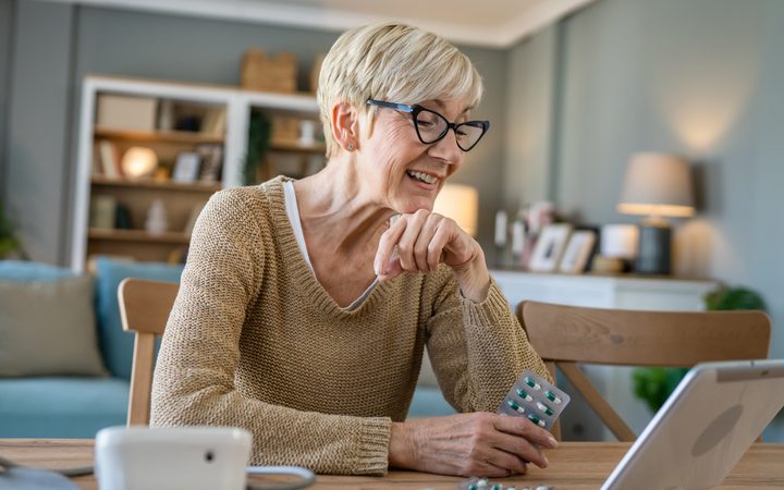 An older woman at home smiling, holding a blister pack beside a tablet computer