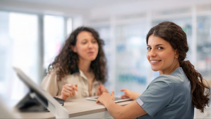 A pharmacy team member at the counter beside a screen, with a customer behind