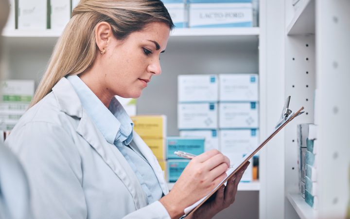 A pharmacist in a white coat writing on a clipboard beside shelves of medicines