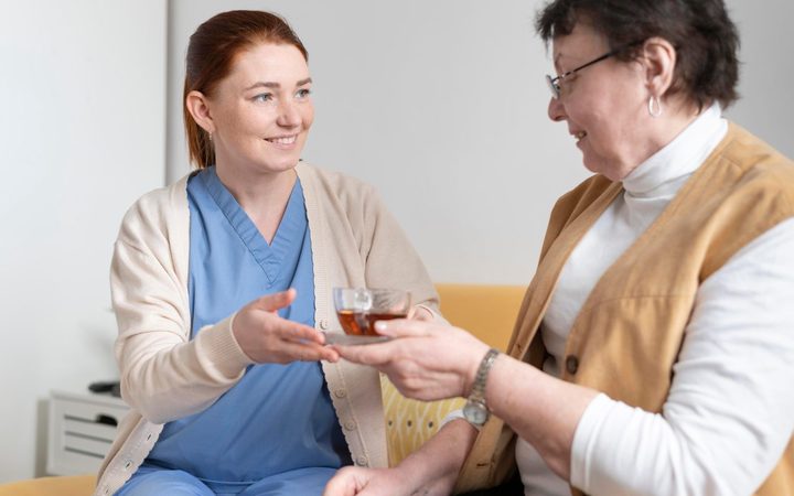 A nurse handing a cup of tea to a patient sitting on a sofa