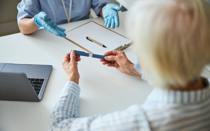A clinician in blue gloves handing an injection pen to an older patient across a desk